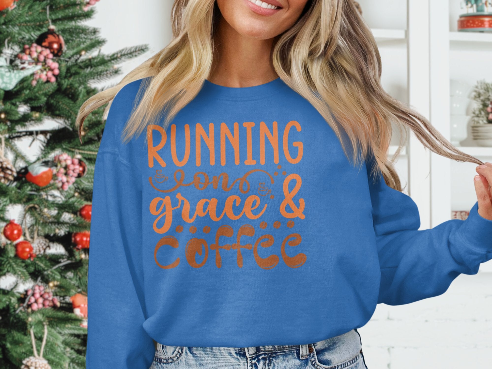 Wearing a cozy, unisex graphic sweatshirt from Garment Graphics that reads Running on grace & coffee, a person stands by an ornamented Christmas tree, surrounded by white shelves and a small plant.