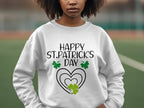 A person with curly hair is outdoors, wearing a festive Garment Graphics sweatshirt featuring a clover design and Happy St. Patricks Day text with shamrocks and hearts, perfectly capturing the spirit of the occasion against a blurred sports field background.