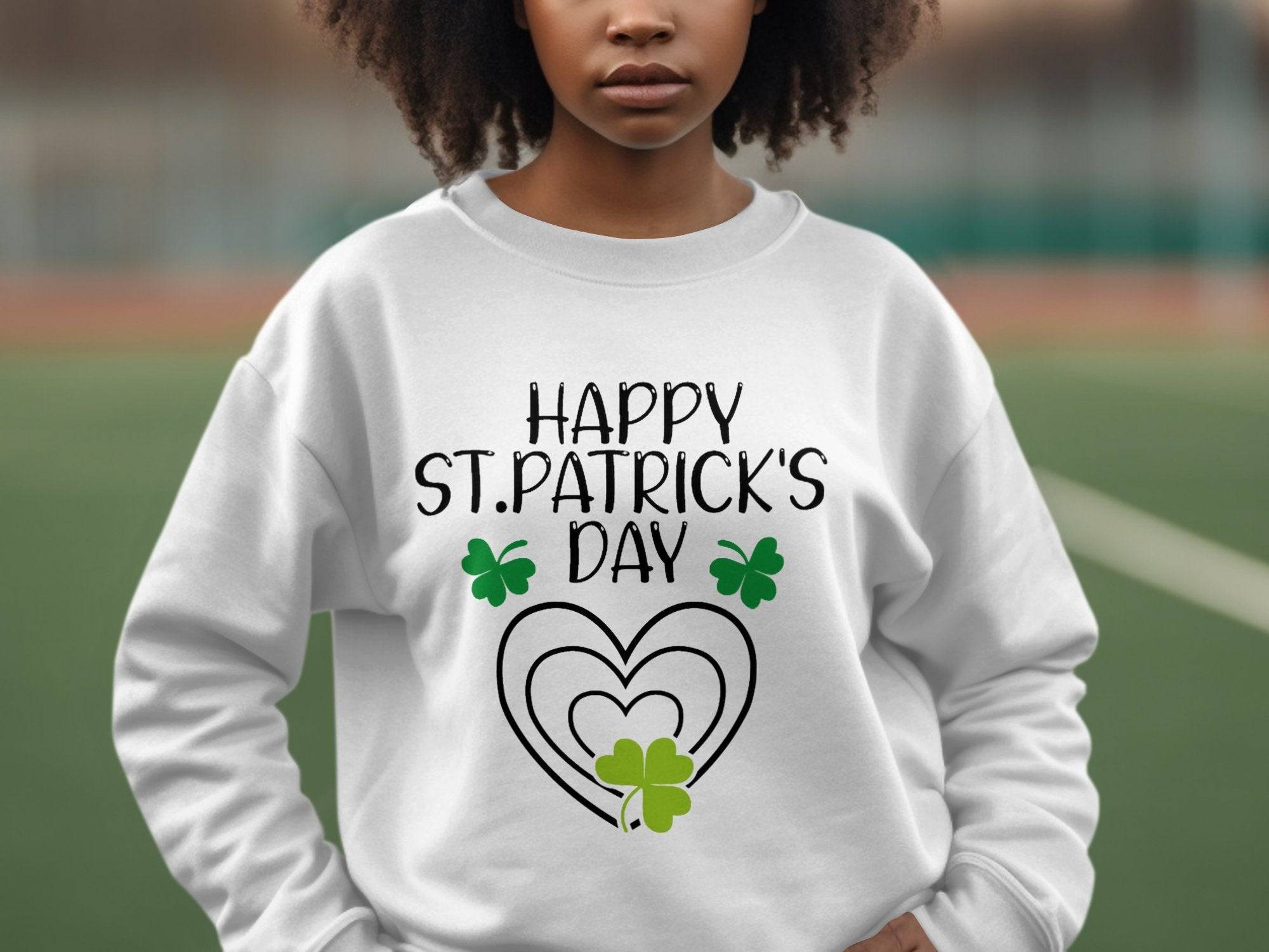 A person with curly hair is outdoors, wearing a festive Garment Graphics sweatshirt featuring a clover design and Happy St. Patricks Day text with shamrocks and hearts, perfectly capturing the spirit of the occasion against a blurred sports field background.