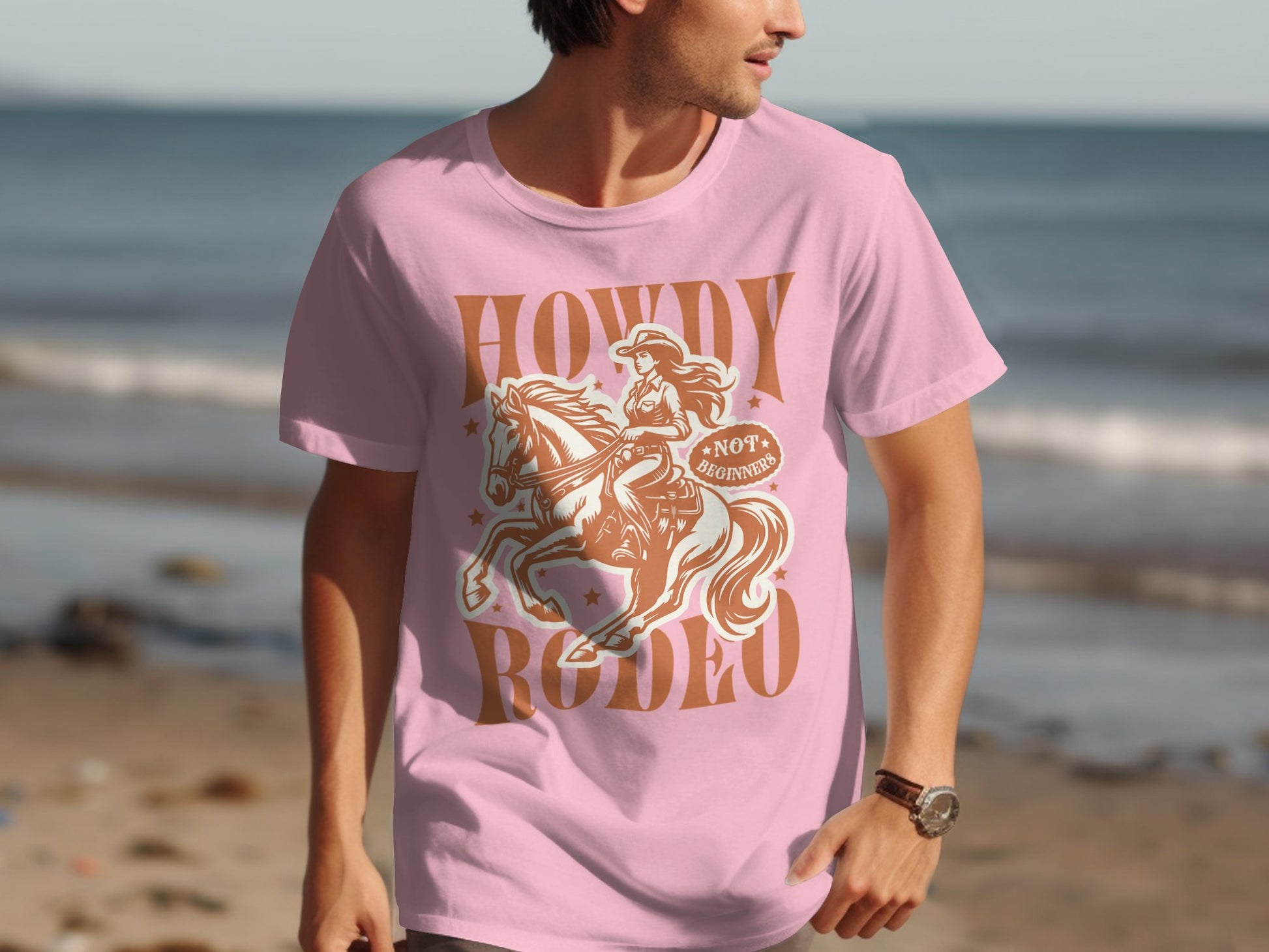 A man wearing a Howdy Rodeo T-shirt by Garment Graphics, adored by rodeo fans, stands on a sandy beach with the ocean behind him; his shirt features a cowboy on a rearing horse.