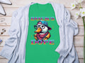 The Garment Graphics green T-shirt boasts a cartoon duck with glasses, hat, and scarf, captioned Ducks Dope. Styled uniquely on a light gray cardigan surrounded by accessories on wood, it becomes an effortless standout piece.