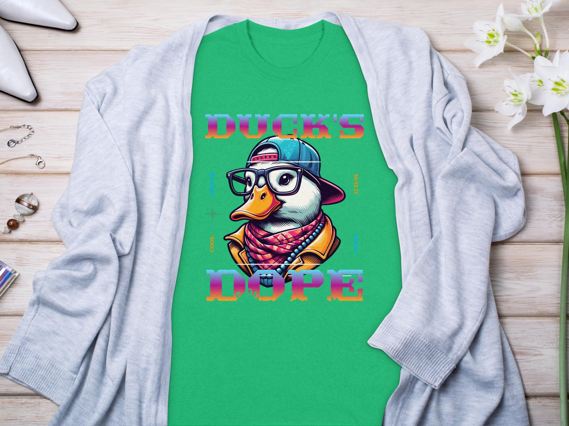 The Garment Graphics green T-shirt boasts a cartoon duck with glasses, hat, and scarf, captioned Ducks Dope. Styled uniquely on a light gray cardigan surrounded by accessories on wood, it becomes an effortless standout piece.