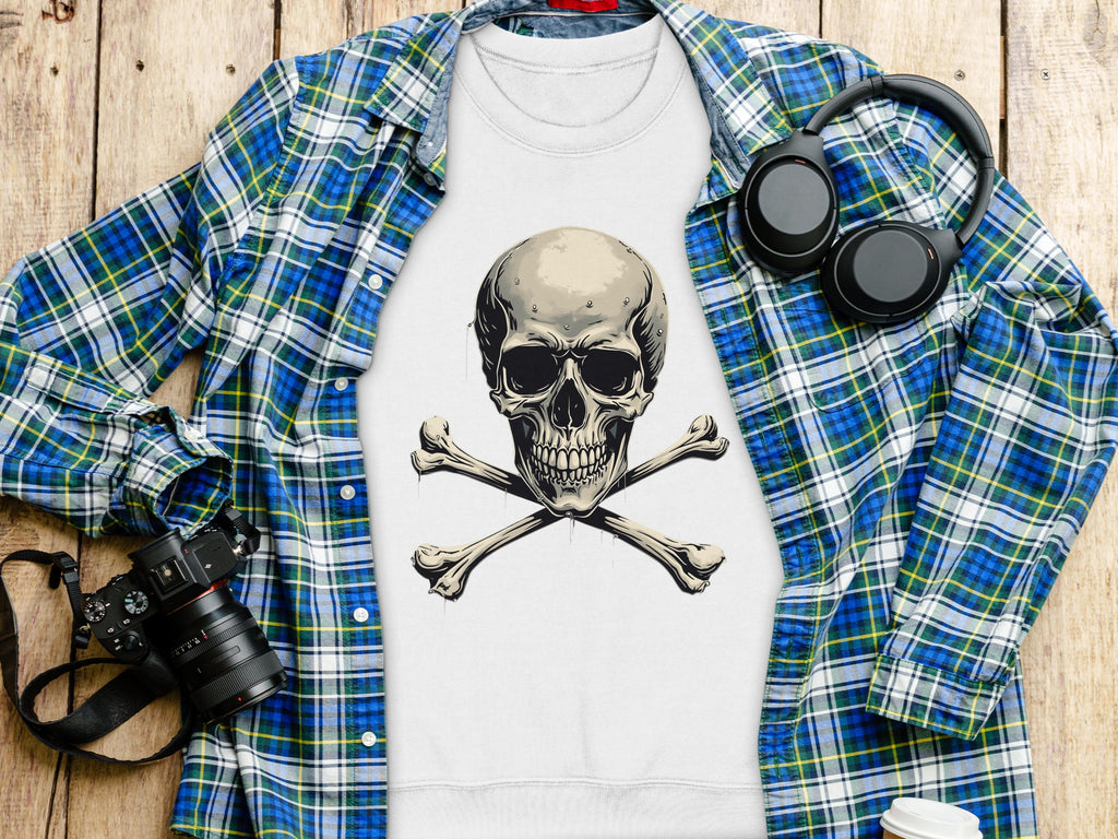 A flat lay on a wooden surface featuring Garment Graphics plaid shirt, a bold white sweatshirt with a skull and crossbones design, black headphones, a camera, notepad, and disposable coffee cup.