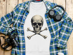 A flat lay on a wooden surface featuring Garment Graphics plaid shirt, a bold white sweatshirt with a skull and crossbones design, black headphones, a camera, notepad, and disposable coffee cup.