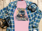 A Garment Graphics T-shirt in pink, featuring a powerful truck and Keep on Trucking, is displayed on a plaid shirt. Nearby are a camera, headphones, and a white cup atop the wooden surface, evoking an adventurous vibe.