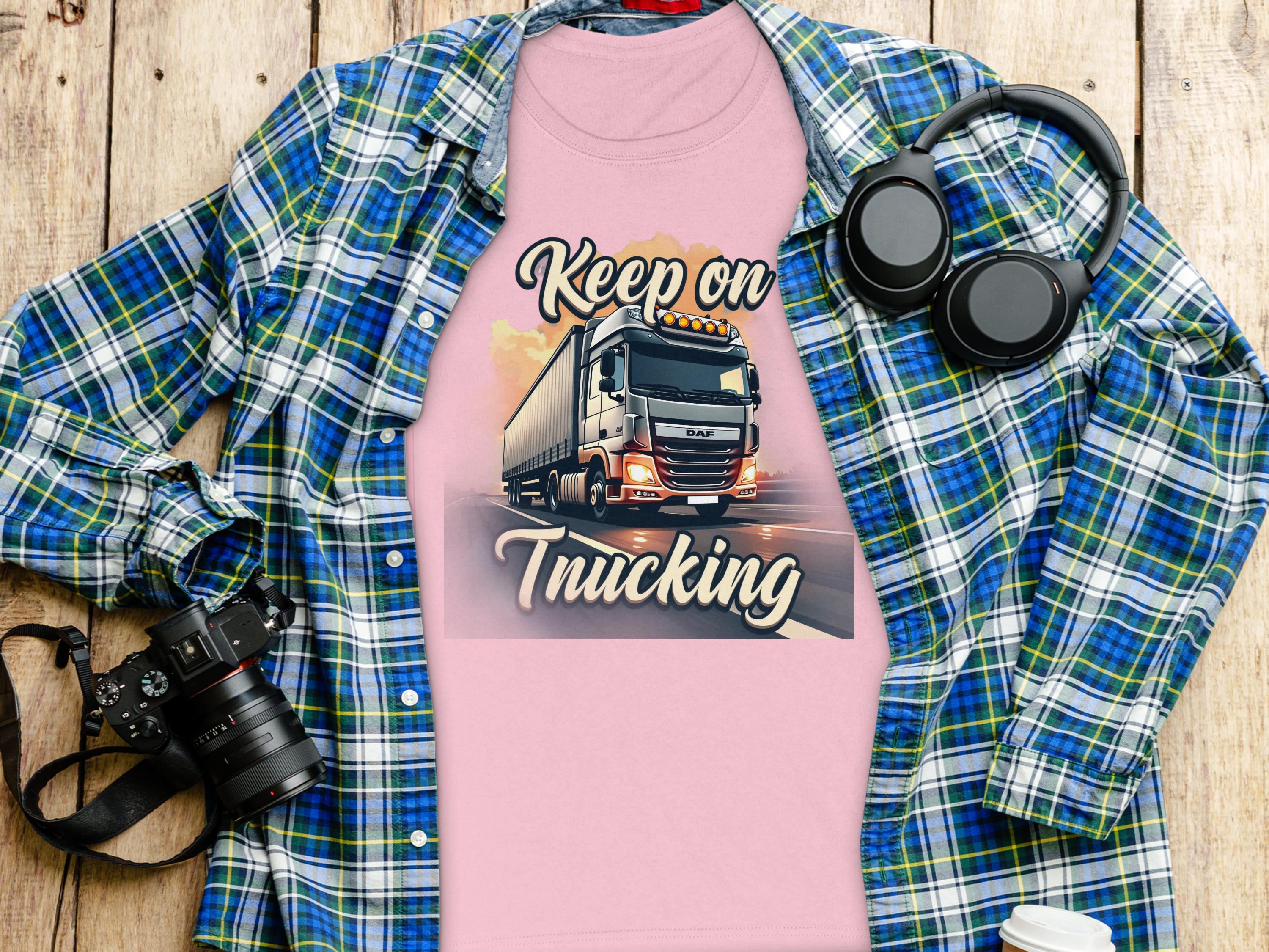 A Garment Graphics T-shirt in pink, featuring a powerful truck and Keep on Trucking, is displayed on a plaid shirt. Nearby are a camera, headphones, and a white cup atop the wooden surface, evoking an adventurous vibe.