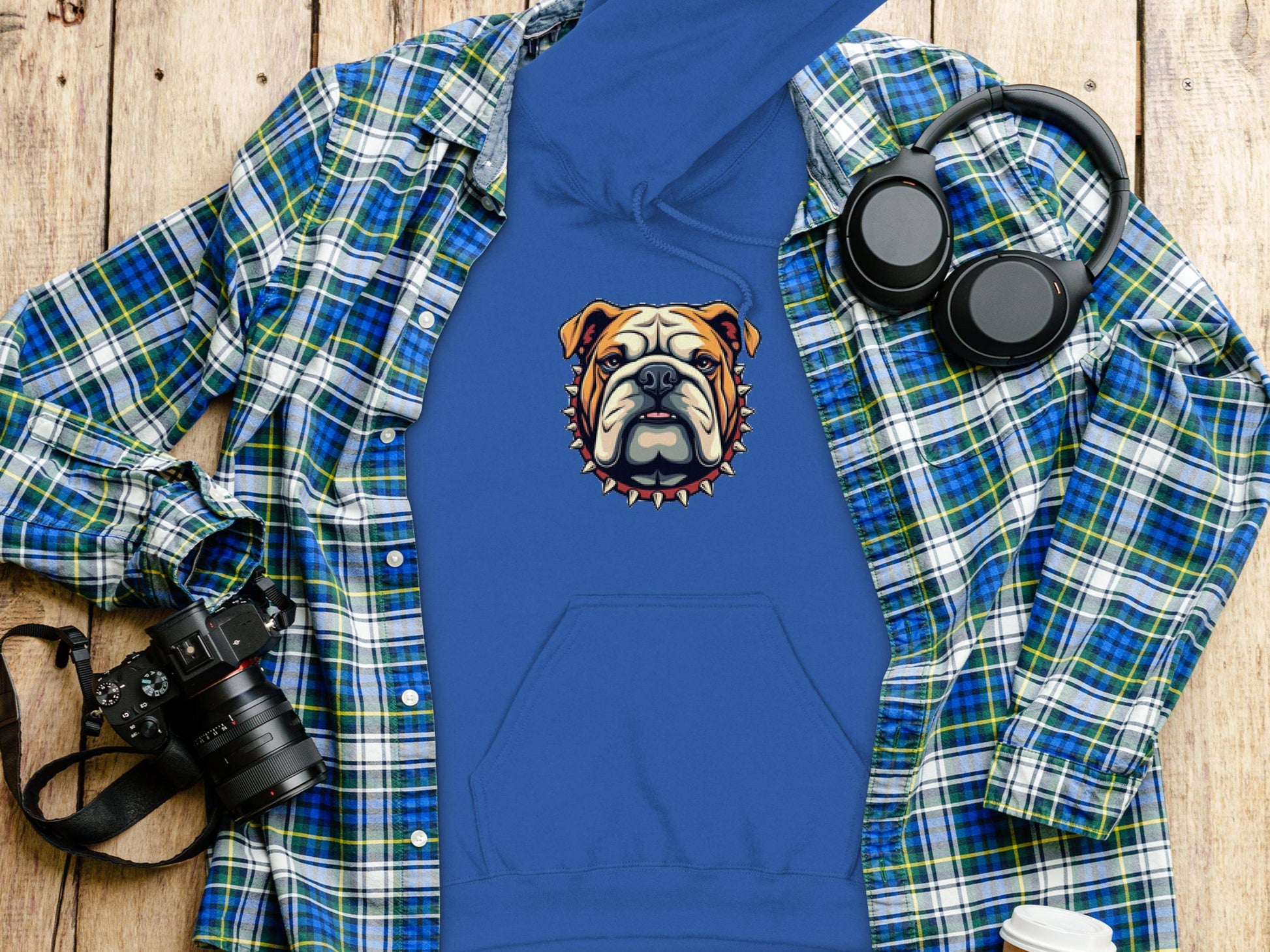 A nostalgic scene features Garment Graphics blue bulldog hoodie with spiked collar on wood, paired with a blue/green plaid shirt, black headphones, a black camera, and a white disposable coffee cup.