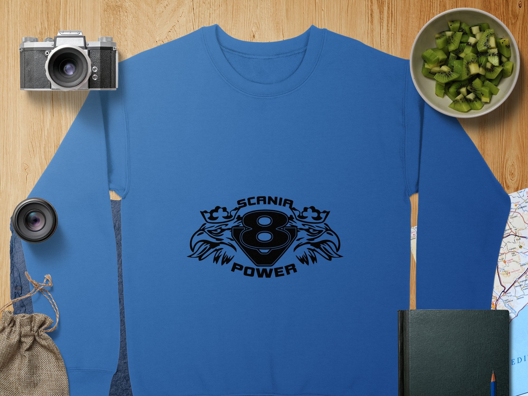 Garment Graphics stylish blue sweatshirt features a Scania Power V8 and eagle in black. Its surrounded by a camera, lens, kiwi slices, small sack, black notebook, and a map on wood—creating an effortlessly cool scene.