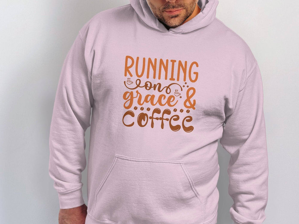 The individual wears a light pink hoodie by Garment Graphics, made from medium-heavy fabric. It features Running on Grace & Coffee in stylish brown text, with a classic fit that stands out against a plain white backdrop.