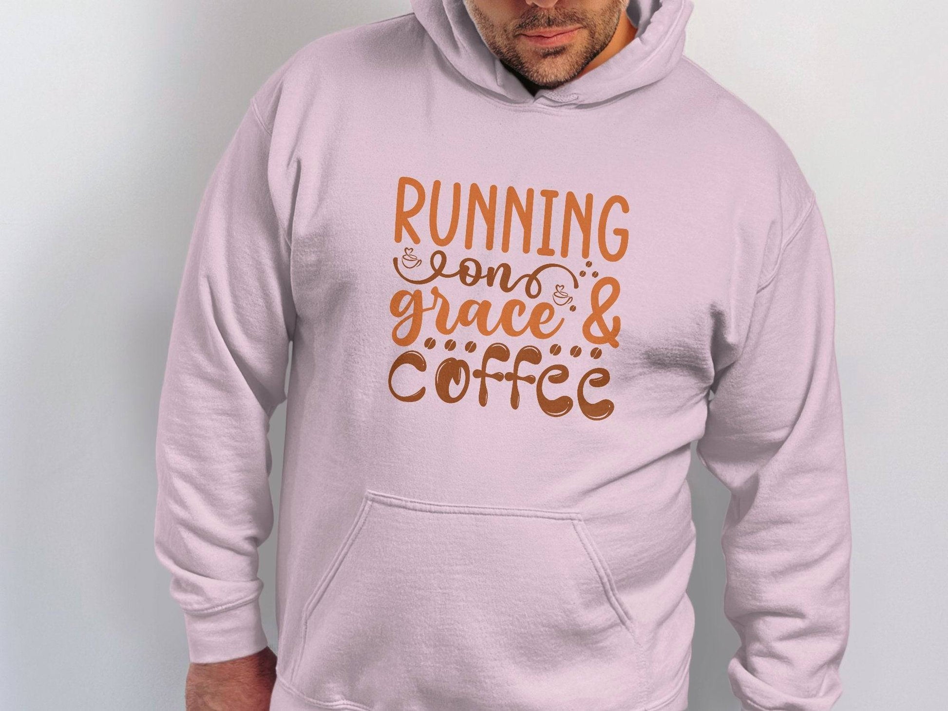 The individual wears a light pink hoodie by Garment Graphics, made from medium-heavy fabric. It features Running on Grace & Coffee in stylish brown text, with a classic fit that stands out against a plain white backdrop.