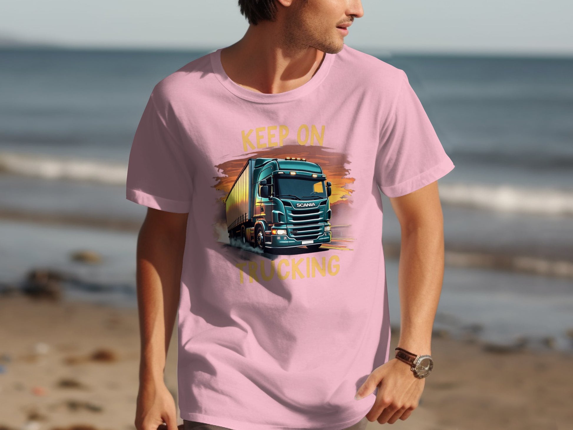 A man at the beach strolls in a pink Garment Graphics T-shirt, featuring a bold truck design and the phrase Keep On Trucking, warmly framed by golden sand and glistening ocean waves.