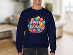 A person wears a navy blue Happy Birthday sweatshirt by Garment Graphics, featuring colorful balloons in unisex sizing. The blurred living room background enhances the cozy celebration vibe.