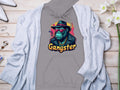 A Garment Graphics gray hoodie showcases a hipster cartoon gorilla in a fedora, sunglasses, and gold chain with Gangster below. Its styled on wood next to a light gray cardigan, white flowers, and accessories for the perfect urban blend.