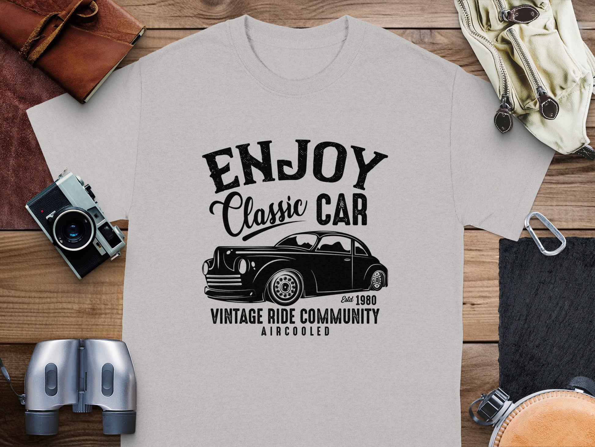 The Garment Graphics gray T-shirt showcases a vintage car graphic and the text Enjoy Classic Car, Vintage Ride Community, Aircooled, Estd. 1980. Ideal for classic car fans, it pairs well with a camera, binoculars, and leather accessories displayed on wood.