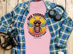 Garment Graphics Bee Yourself T-Shirt features a cartoon bee with sunglasses and a crown, surrounded by Bee Yourself, Everyone Else is Taken, Summer Edition! Displayed on wood with a plaid shirt, camera, headphones, and coffee; now available in womens sizes.