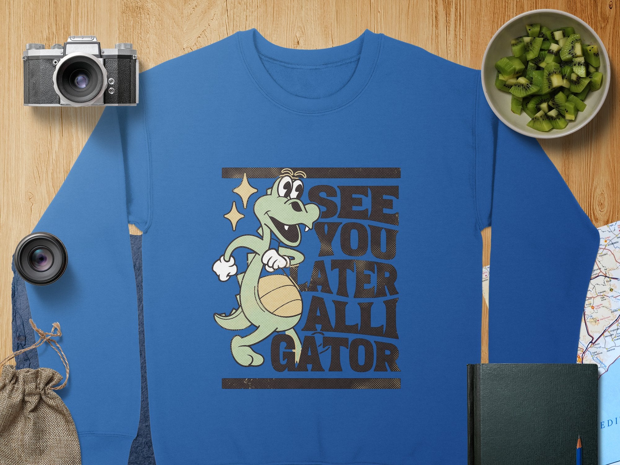 A blue Garment Graphics sweatshirt with a playful alligator graphic and See You Later Alligator text is on a wooden surface. Nearby are a camera, lens, bowl of chopped kiwi, burlap pouch, black book, and part of a map.