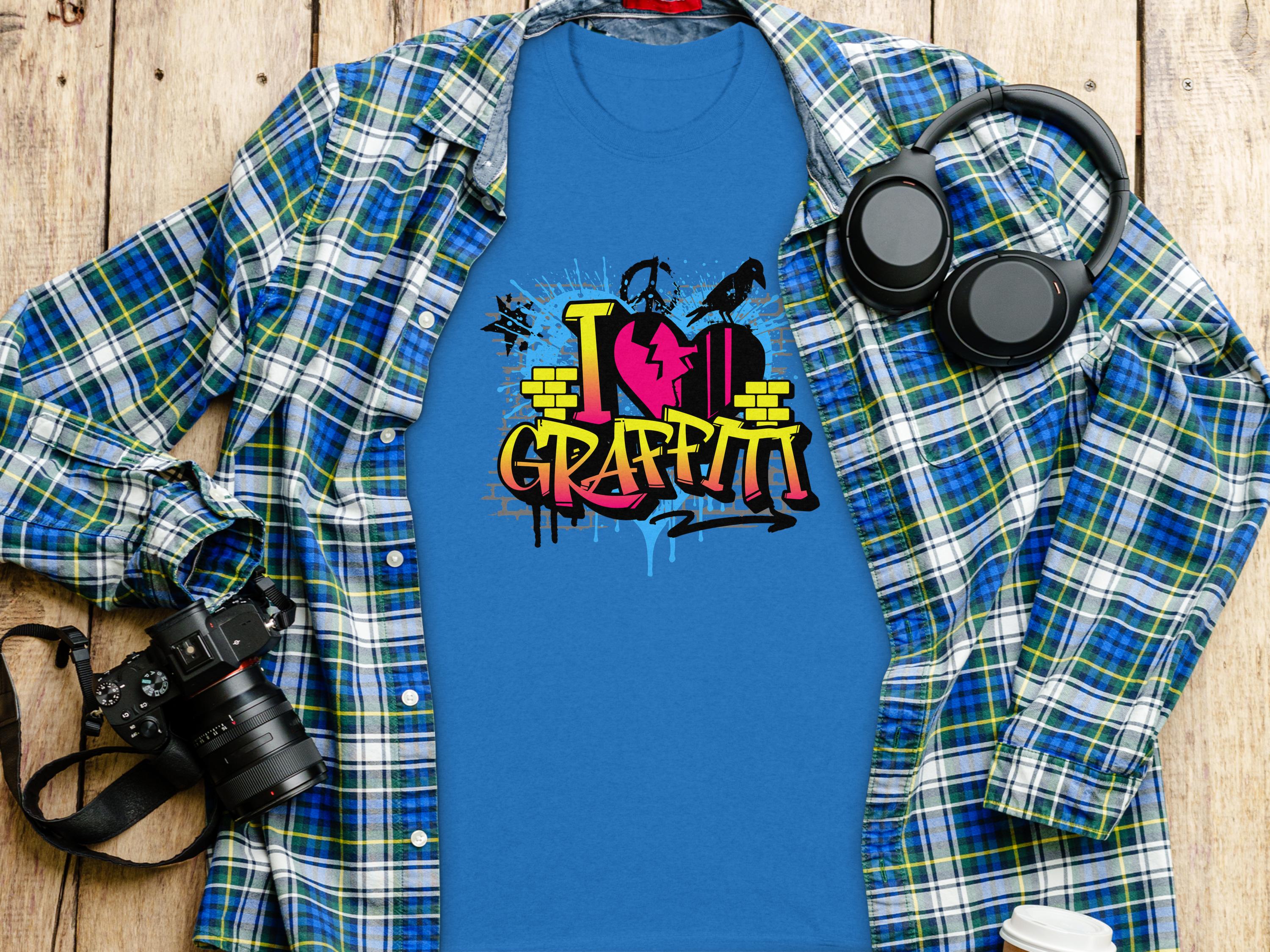 A blue Graffiti Love T-Shirts by Garment Graphics, featuring a vibrant street art design with I Love Graffiti, lies on a wooden surface beneath a blue and green plaid shirt. Nearby are over-ear headphones, a camera, and a notebook, creating an artistic ensemble.