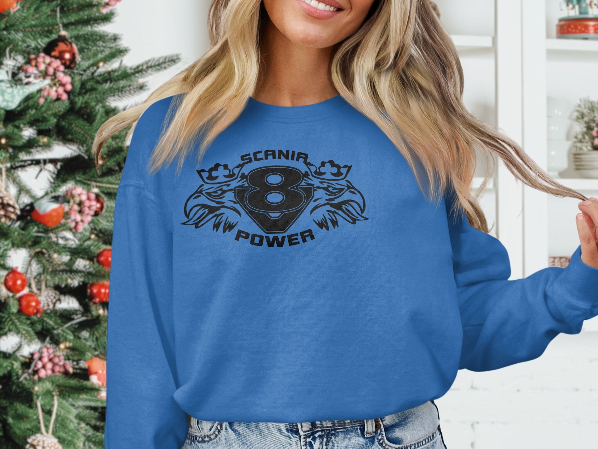 A person in a Garment Graphics blue sweater with the 8 Scanir Power design and two stylized leopards stands by a decorated Christmas tree, twirling their blonde hair, while white shelves with decor are seen in the background.