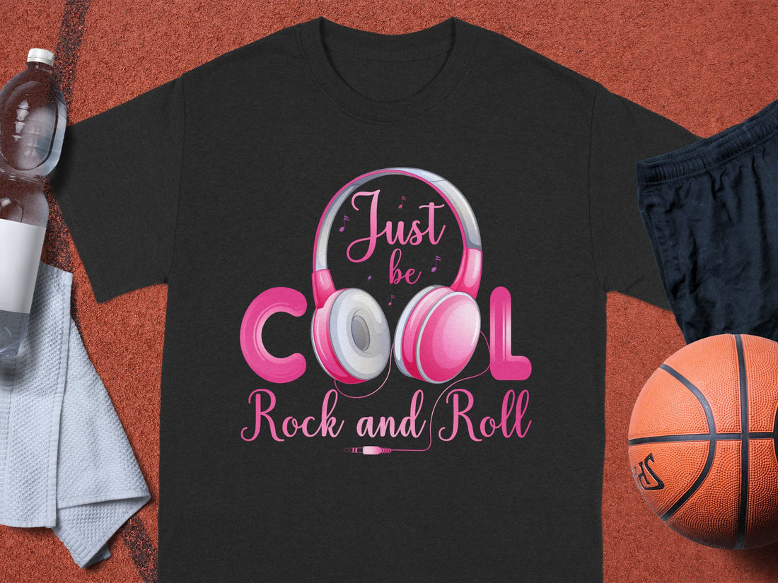 A music enthusiast wears a Garment Graphics white T-shirt with pink Fun Headphones and the text Just be Cool, Rock and Roll, standing outdoors in sand with tall grass swaying behind them.