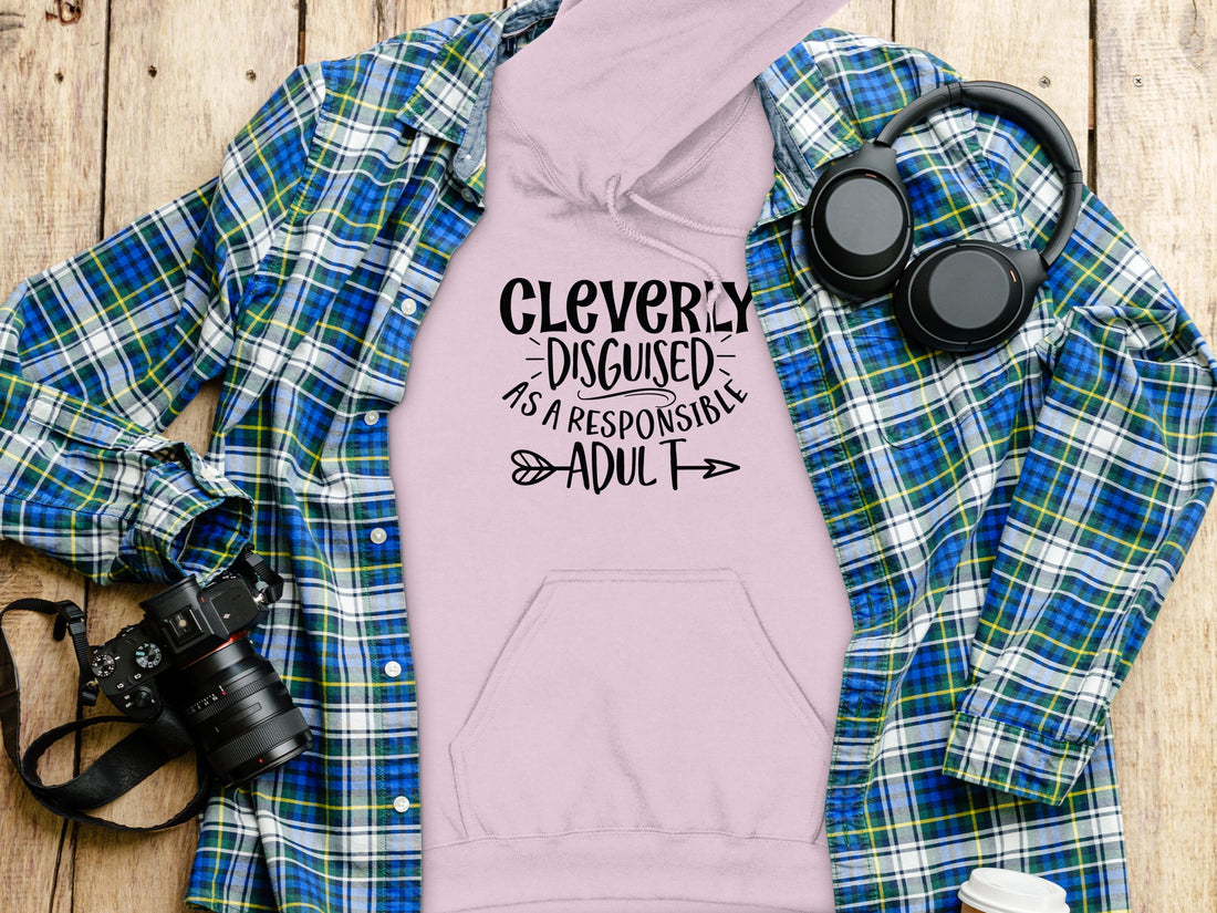 A person wears a black hoodie from Garment Graphics Cleverly Disguised Hoodies collection, featuring Cleverly Disguised As A Responsible Adult on the front. The plain white background highlights this casual wear staple.