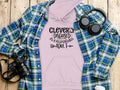 A lavender hoodie from Garment Graphics Responsible Adult Hoodies collection, featuring the text Cleverly disguised as a responsible adult, is displayed on wood, surrounded by a blue plaid shirt, headphones, a camera, and a travel mug.