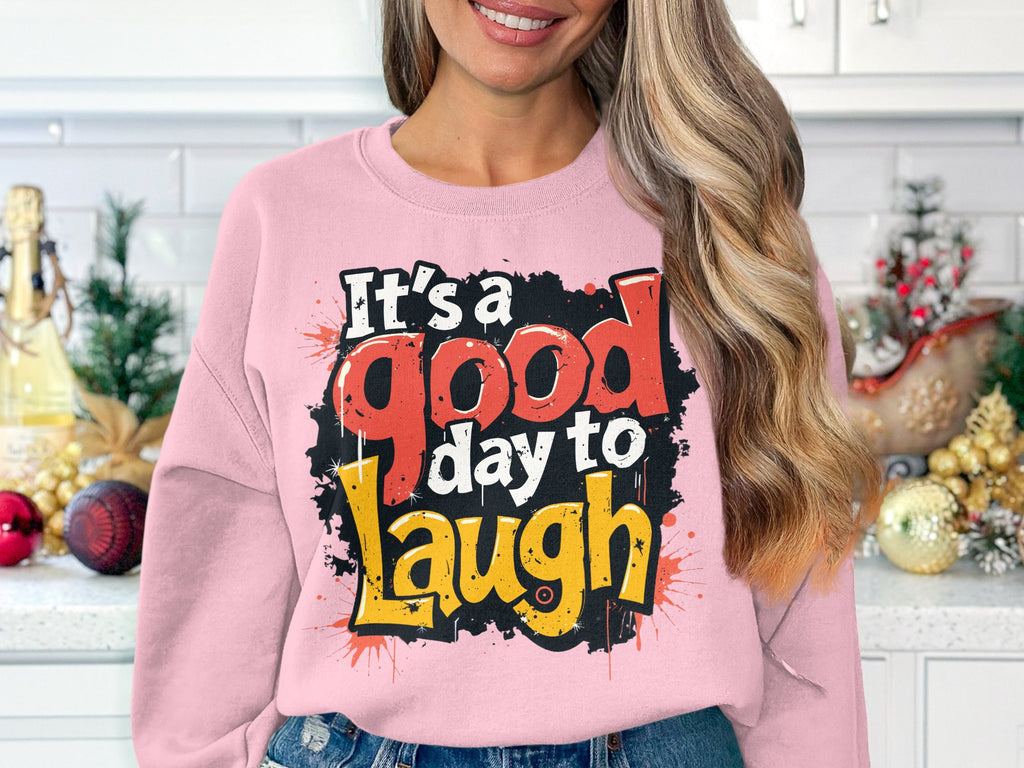 In a kitchen with festive decorations and white cabinets, someone wears a medium-heavy fabric pink sweatshirt by Garment Graphics. The unisex sweatshirt features Its a good day to laugh boldly printed.