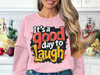 In a kitchen with festive decorations and white cabinets, someone wears a medium-heavy fabric pink sweatshirt by Garment Graphics. The unisex sweatshirt features Its a good day to laugh boldly printed.
