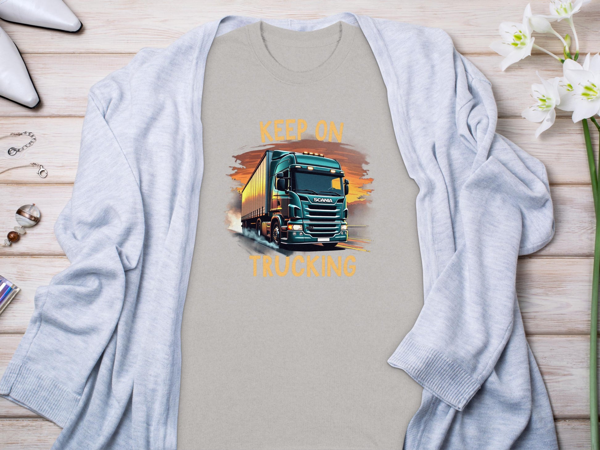 The Garment Graphics gray graphic T-shirt for truck enthusiasts showcases a large truck and KEEP ON TRUCKING text. Its displayed on a wooden surface with jewelry, high heels, white flowers, and framed by a cozy gray cardigan.