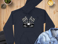 A Garment Graphics black hoodie with a race car and checkered flags graphic lies stylishly on wood, paired with a denim jacket, latte, and potted plant—ideal for motorsport fans who love casual flair.