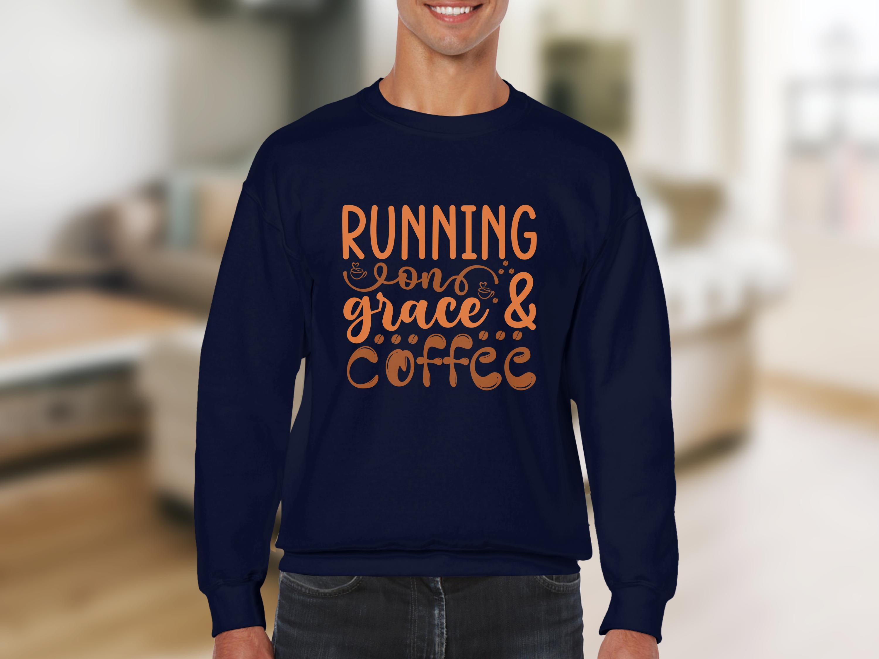 Indoors, a person wears a Garment Graphics navy blue graphic sweatshirt featuring the phrase Running on grace & coffee in vibrant orange. The unisex sizing offers comfort for all, set against a blurred backdrop of neutral-toned decor.
