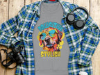 A Garment Graphics gray T-shirt with a vibrant dog in sunglasses and PAWSITIVE STYLISH text rests on wood. Nearby: a blue plaid shirt, black headphones, a camera, and a white cup with lid—ideal for dog lovers.