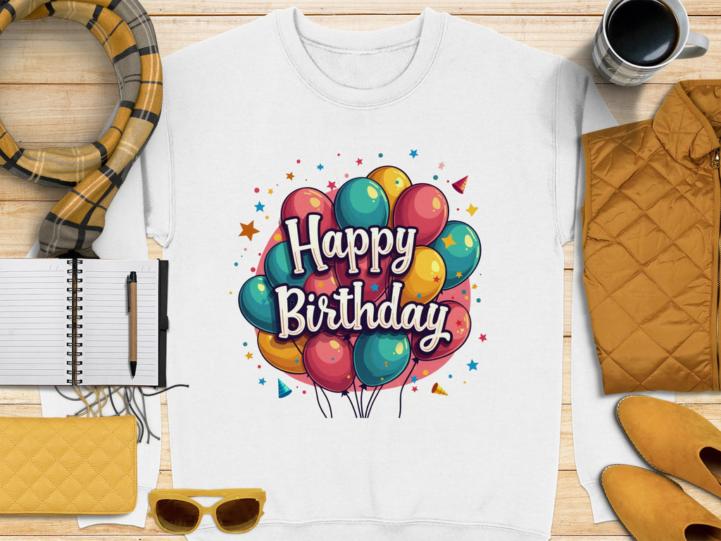 A flat lay highlights a Garment Graphics unisex white Happy Birthday sweatshirt with colorful balloons, accompanied by a scarf, notebook with pen, yellow clutch, sunglasses, orange jacket, and a coffee cup on wooden surface.