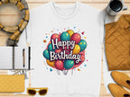 A flat lay highlights a Garment Graphics unisex white Happy Birthday sweatshirt with colorful balloons, accompanied by a scarf, notebook with pen, yellow clutch, sunglasses, orange jacket, and a coffee cup on wooden surface.