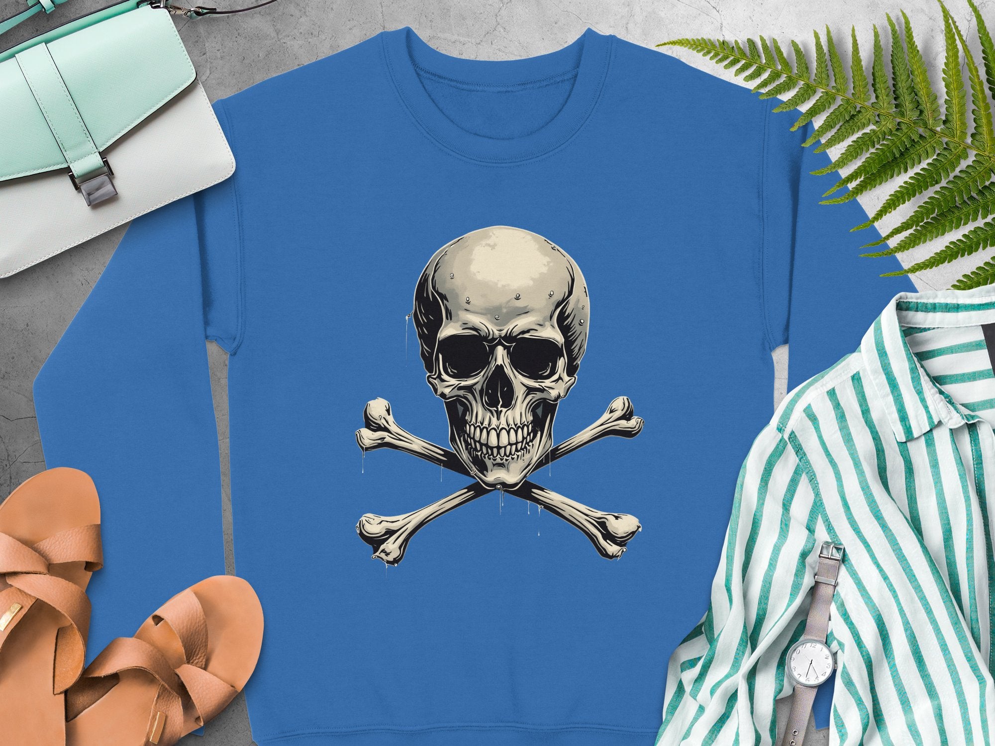 Garment Graphics offers a blue sweatshirt with a skull and crossbones graphic for an edgy look. It pairs well with a mint handbag, tan sandals, striped shirt, wristwatch, and fern leaf on a light gray surface. Unisex sizing ensures versatile wear for all styles.