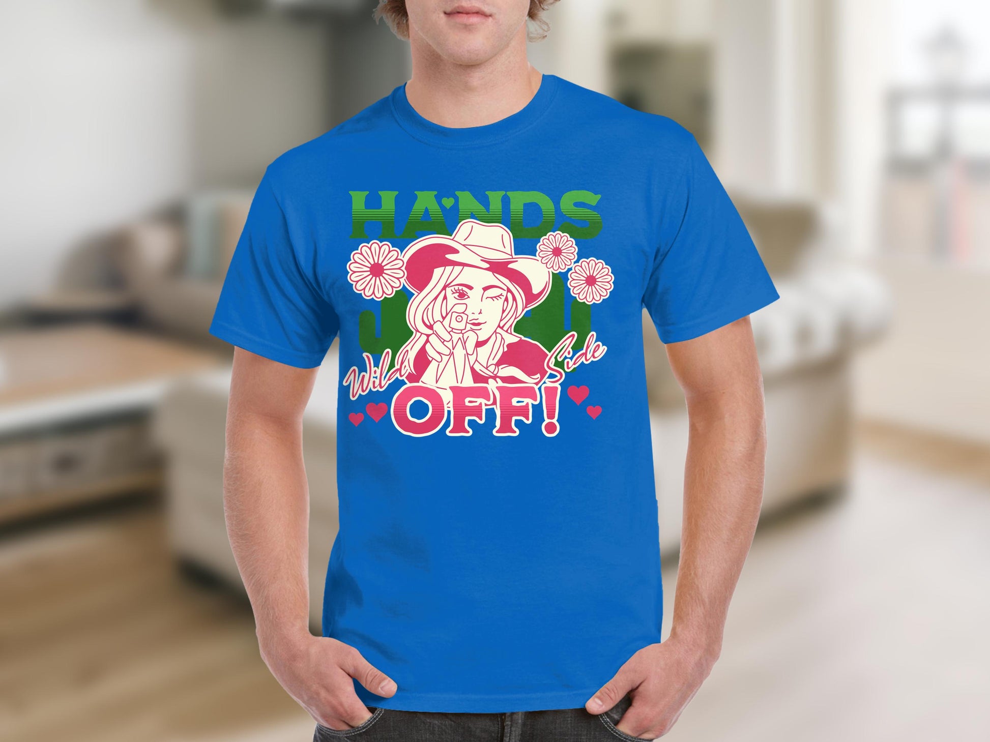 Someone wears a Garment Graphics classic fit blue t-shirt with HANDS OFF! and a bold cowboy hat, lasso, flowers, and hearts design. The indoor background features a cozy couch.
