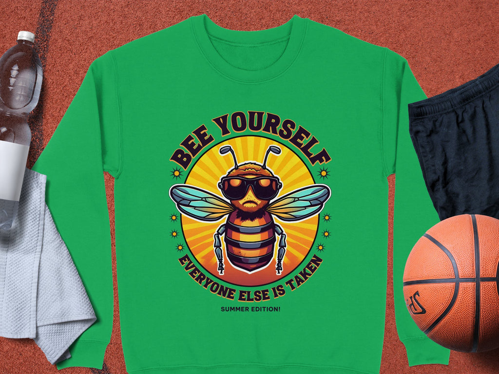 Garment Graphics green unisex sweatshirt, featuring a cartoon bee in sunglasses and jacket with Bee Yourself, Everyone Else Is Taken, Summer Edition! text. Styled with a basketball, black shorts, gray towel, and water bottle. Ideal for those embracing individuality!.