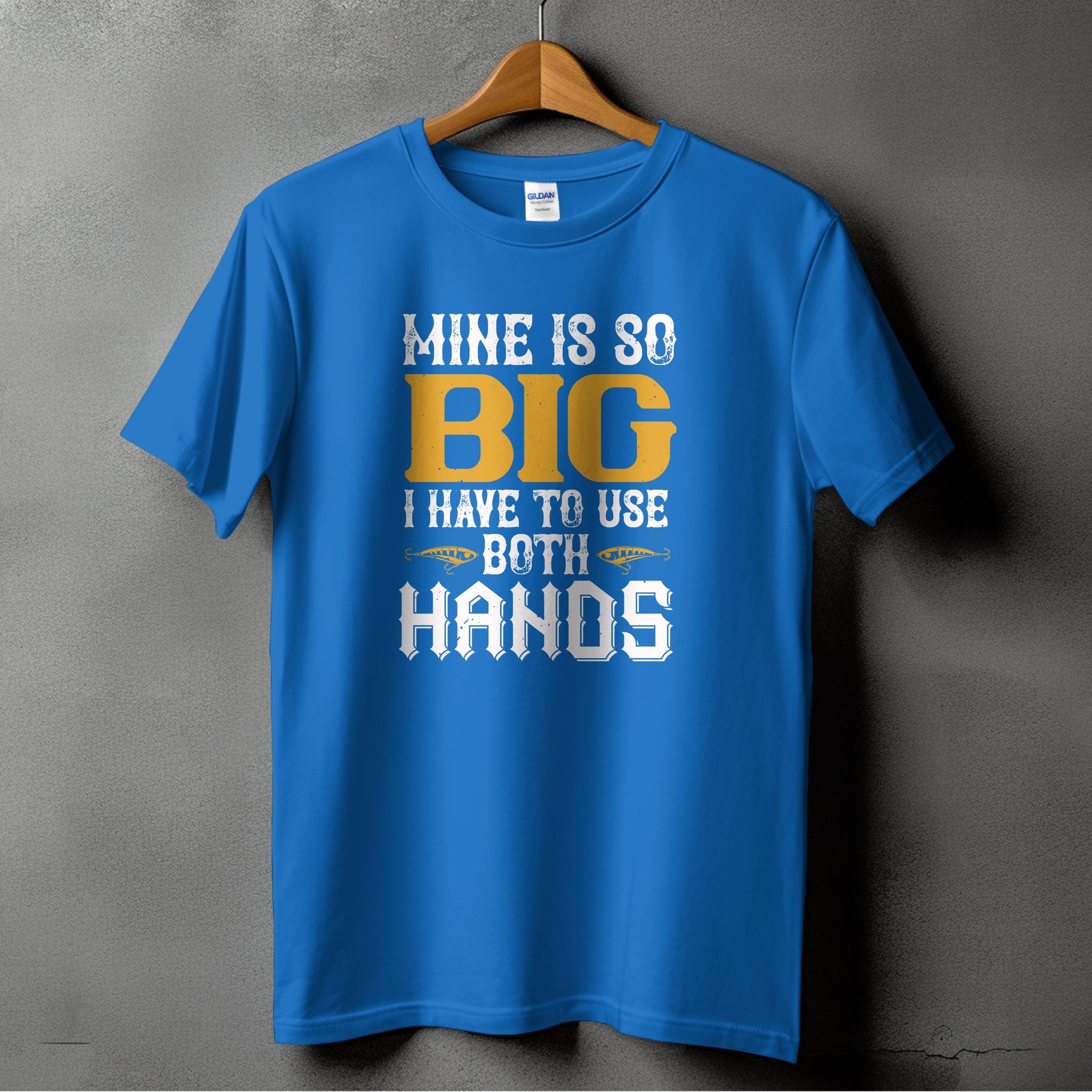 A blue T-shirt from Garment Graphics hangs on a wooden hanger against a gray wall. This fishing-themed tee humorously displays MINE IS SO BIG I HAVE TO USE BOTH HANDS in white and yellow, ideal for fishing enthusiasts with a sense of humor.