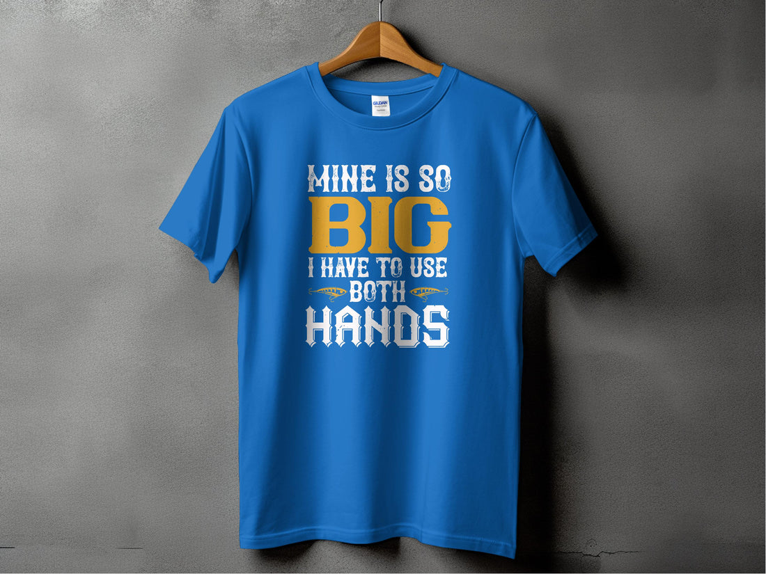 A blue T-shirt from Garment Graphics hangs on a wooden hanger against a gray wall. This fishing-themed tee humorously displays MINE IS SO BIG I HAVE TO USE BOTH HANDS in white and yellow, ideal for fishing enthusiasts with a sense of humor.