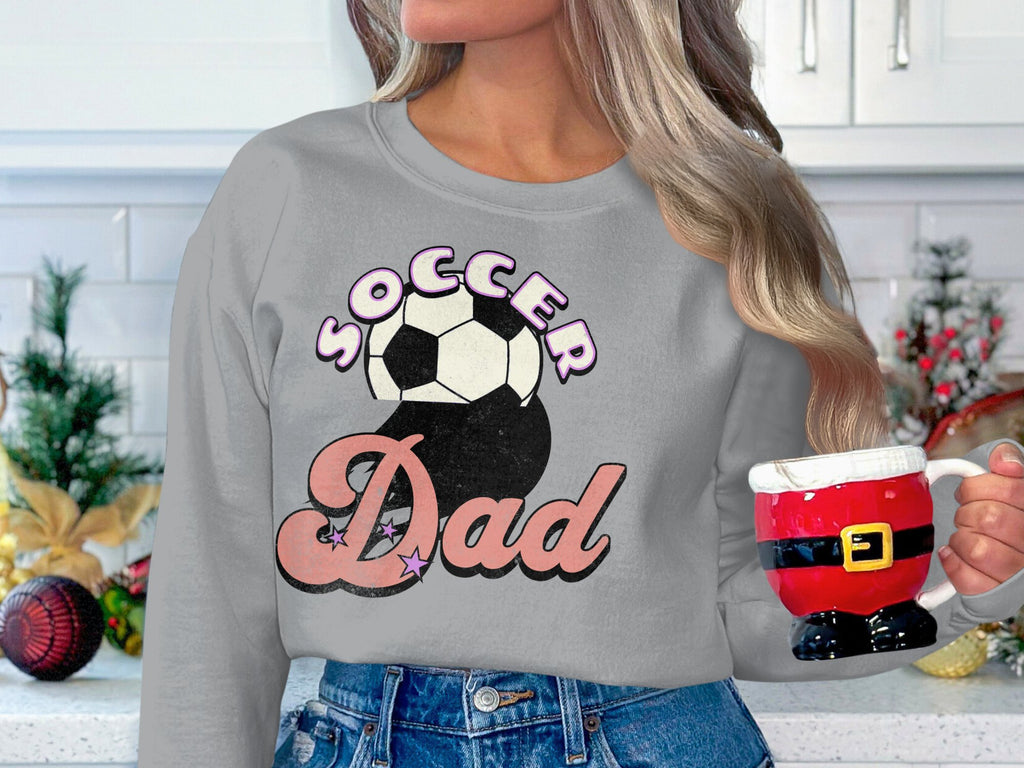 In a festively decorated kitchen with a plant, a long-haired person holds a Santa-themed mug, donning a Garment Graphics sweatshirt with a retro Soccer Dad design featuring bold and script font soccer balls.