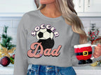 In a festively decorated kitchen with a plant, a long-haired person holds a Santa-themed mug, donning a Garment Graphics sweatshirt with a retro Soccer Dad design featuring bold and script font soccer balls.