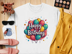 A flat lay image features a white Happy Birthday sweatshirt from Garment Graphics, ideal for celebrations, in unisex sizing. The surrounding items include a yellow jacket, pink shoes, sunglasses, makeup brushes, an eyeshadow palette, nail polish, and a drawstring bag on a wooden surface.