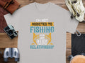 A Garment Graphics T-shirt with the design Im not addicted to fishing. We are just in a very committed relationship. is displayed on wooden surface surrounded by fishing gear, camera, binoculars, rope, and a leather pouch—ideal for passionate anglers.