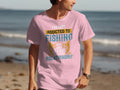 A beachgoer sports a pink Fishing T-shirt by Garment Graphics, featuring the slogan Im not addicted to fishing. We are just in a very committed relationship along with angler-themed graphics, set against an ocean backdrop.