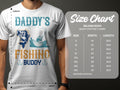 A person wears a Garment Graphics white t-shirt featuring Daddys Fishing Buddy with a fish, boat, and hooks. Nearby is a size chart for Gildan 5000 heavy cotton t-shirts listing five sizes—ideal for fishing fans.