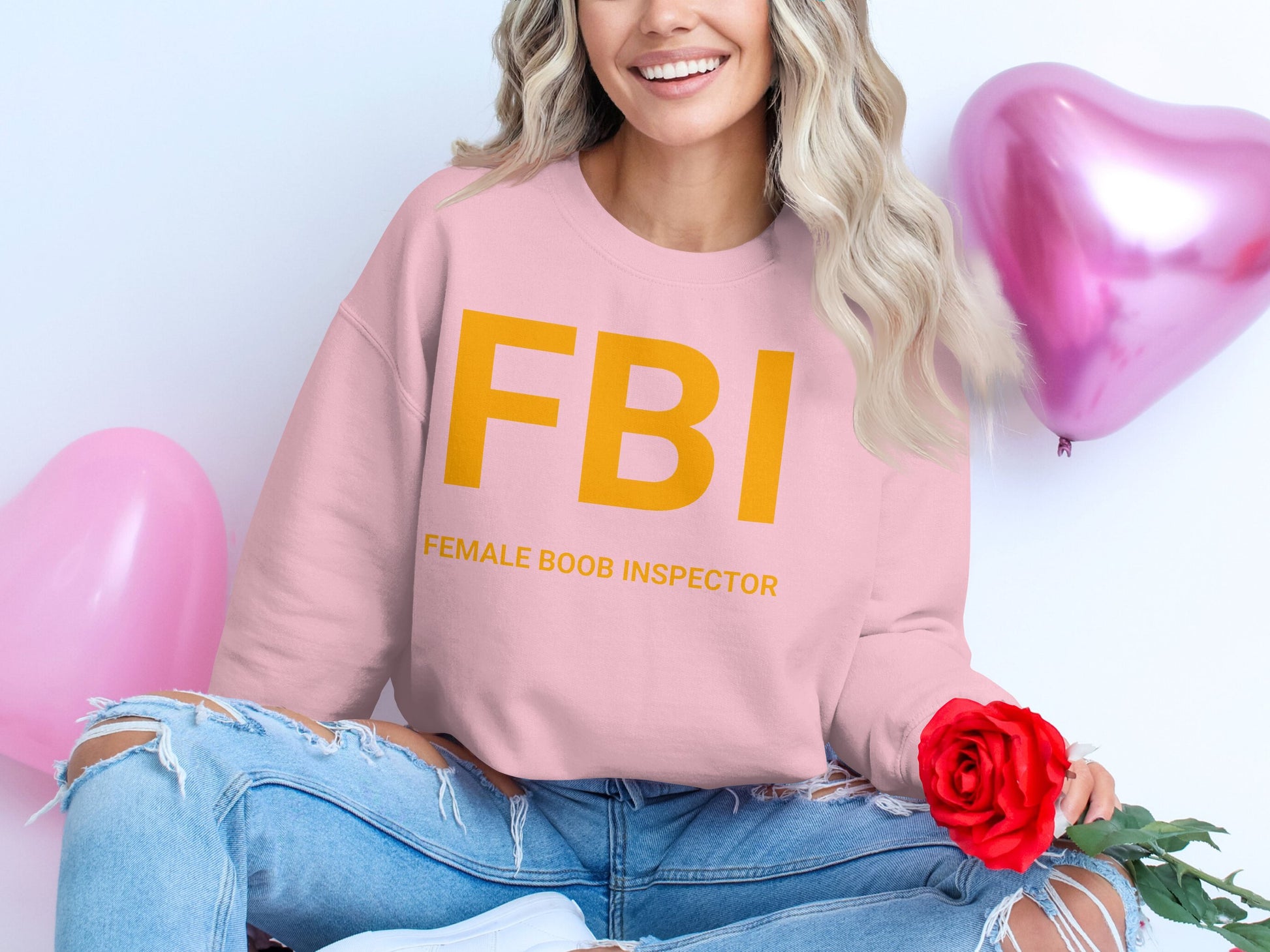 A person with wavy hair sits cross-legged, holding a red rose and smiling. Theyre wearing a Garment Graphics unisex sweatshirt featuring FBI Female Boob Inspector in bold yellow letters. Two pink heart-shaped balloons float behind them, enhancing the playful scene.