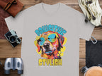 A light gray Dog T-Shirt from Garment Graphics features a dog in sunglasses, with PAWSITIVE above and STYLISH below. Titled Chillin Like A Canine Villain, its displayed on wood with a camera, binoculars, and gloves nearby.
