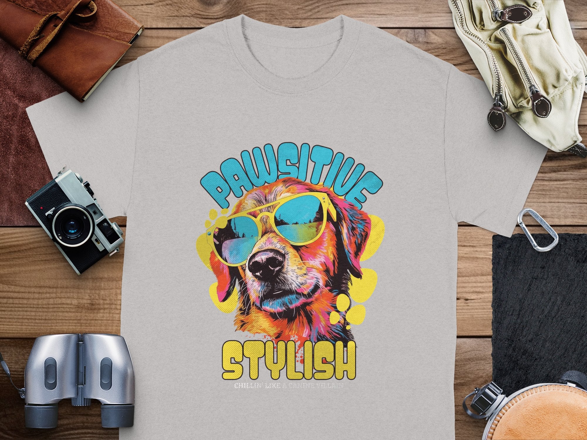 A light gray Dog T-Shirt from Garment Graphics features a dog in sunglasses, with PAWSITIVE above and STYLISH below. Titled Chillin Like A Canine Villain, its displayed on wood with a camera, binoculars, and gloves nearby.
