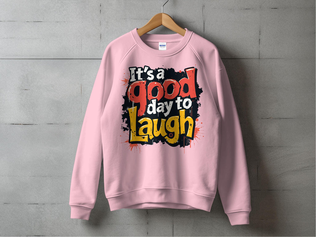 A pink Garment Graphics sweatshirt hangs on a wooden hanger against a gray wall, showcasing the creative flair of graphic art sweatshirts. It features the text Its a Good Day to Laugh in bold, colorful fonts with a splattered paint effect. This cozy piece is perfect for everyone with unisex sizing.