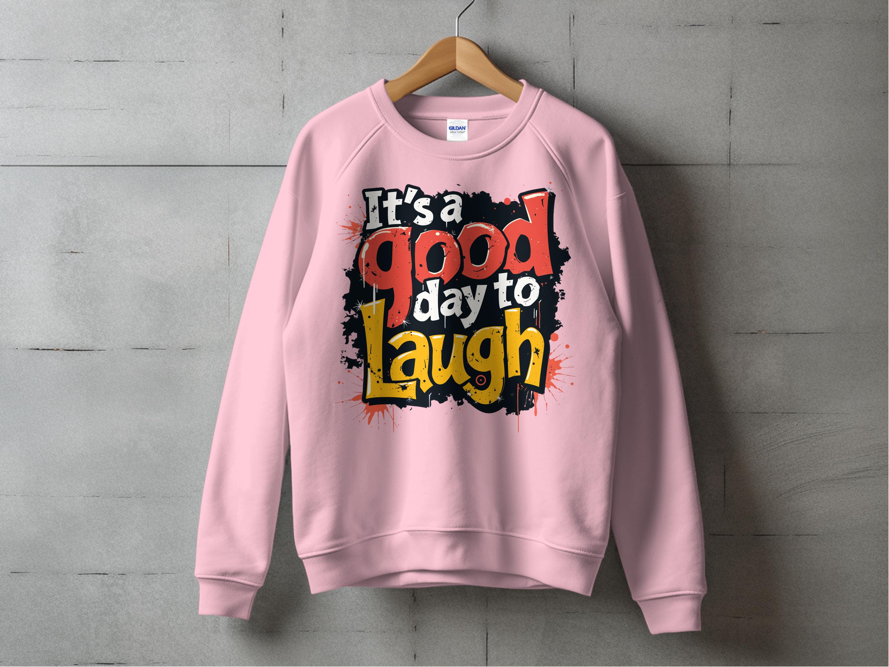 A pink Garment Graphics sweatshirt hangs on a wooden hanger against a gray wall, showcasing the creative flair of graphic art sweatshirts. It features the text Its a Good Day to Laugh in bold, colorful fonts with a splattered paint effect. This cozy piece is perfect for everyone with unisex sizing.