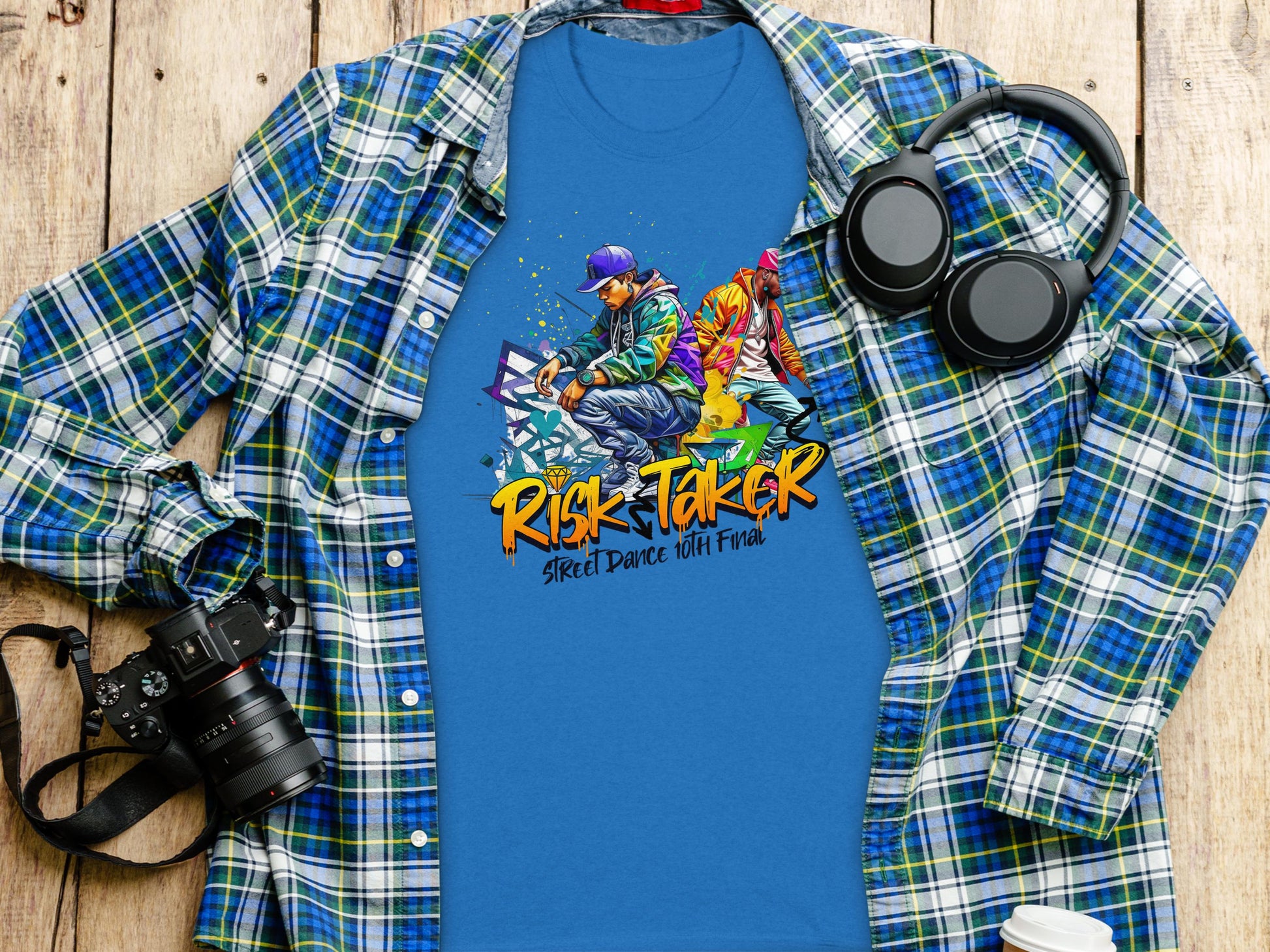 Garment Graphics blue T-shirt, with graffiti-style Risk Taker Street Dance 10th Final design, is laid on wood beside a plaid shirt, headphones, camera, and watch—embodying street dance culture.